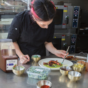 A chef is plating a dish of octopus with green sauce and garnishes using a pair of tongs to arrange the octopus.