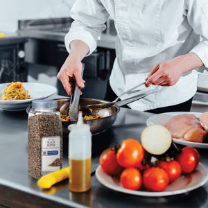 A chef is using tongs to stir a pan of food while a jar of McCormick Culinary Cracked Black Pepper sits on the counter.