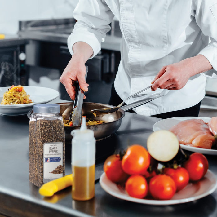 A chef is using tongs to stir a pan of food while a jar of McCormick Culinary Cracked Black Pepper sits on the counter.