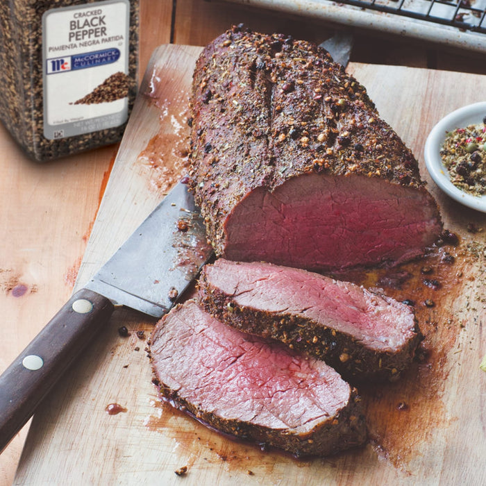 A close-up of a sliced roast beef seasoned with McCormick Culinary Cracked Black Pepper on a wooden cutting board.