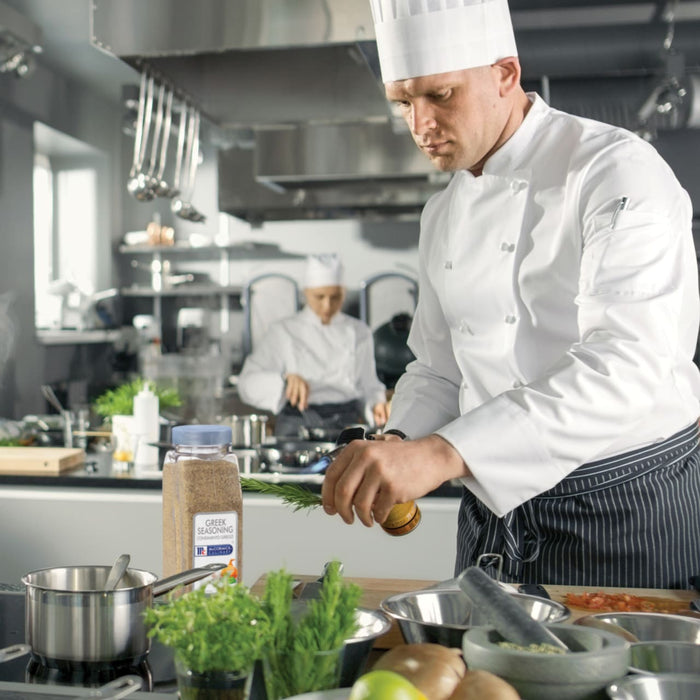 A chef in a white chefs coat is using a blowtorch to cook a dish with a jar of McCormick Culinary Greek Seasoning in the background.