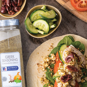 A bottle of McCormick Culinary Greek Seasoning next to a pita bread filled with chicken tomatoes cucumbers spinach feta cheese and olives.