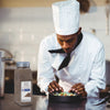 A chef in a white uniform is plating a dish of food while holding a shaker of McCormick Culinary Black Pepper.