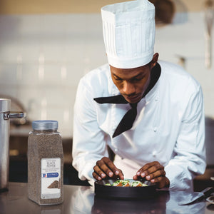 A chef in a white uniform is plating a dish of food while holding a shaker of McCormick Culinary Black Pepper.