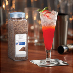 A glass of red cocktail with a basil garnish sits on a black and white napkin next to a McCormick Culinary Black Pepper shaker.