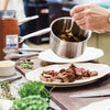 A plate of sliced steak with a side of herbs and a pot of barbecue sauce.