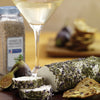 A close-up of a log of goat cheese covered in herbs de Provence with a glass of white wine and figs in the background.