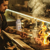 A chef uses a pair of tongs to flip a piece of meat on a hot grill with a bottle of McCormick Grill Mating in the background.
