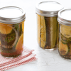 Three glass jars of dill pickles two with lids on and one with the lid off sitting on a white table with a red and white striped napkin.