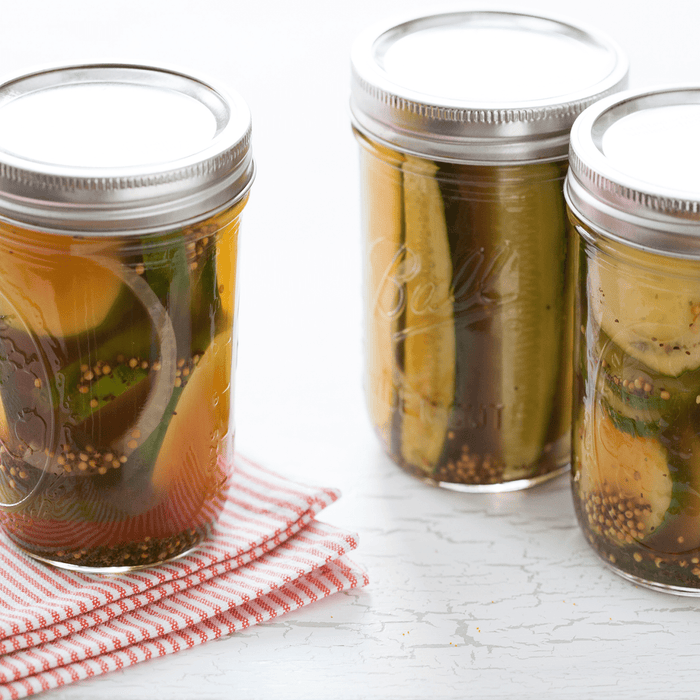 Three glass jars of dill pickles two with lids on and one with the lid off sitting on a white table with a red and white striped napkin.