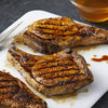 Three grilled pork chops with grill marks are arranged on a white cutting board with a bowl of sauce and a fork.