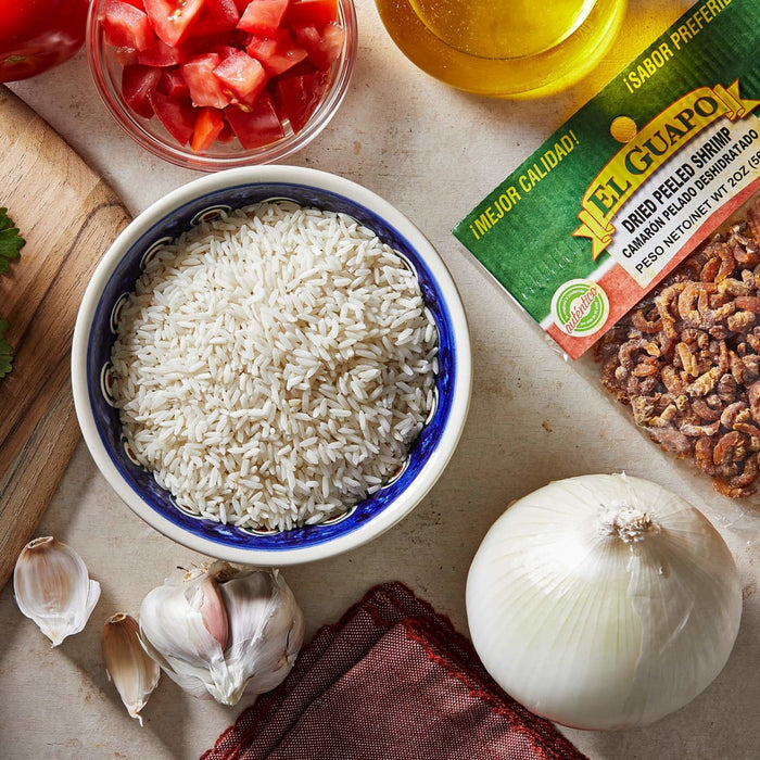 A bowl of white rice a bag of El Guapo dried peeled shrimp garlic a white onion and diced tomatoes.