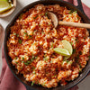 A close-up of a skillet filled with red rice cilantro and lime wedges.