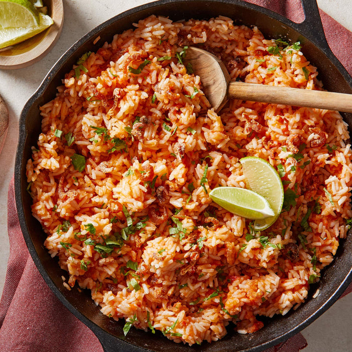A close-up of a skillet filled with red rice cilantro and lime wedges.