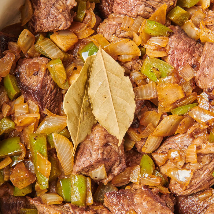 A close-up of a dish of beef stew with onions, peppers, and bay leaves.