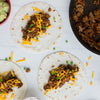 Three tortillas are being prepared with seasoned ground beef, shredded cheese, and chopped green onions, with a bowl of guacamole and a skillet of ground beef nearby.