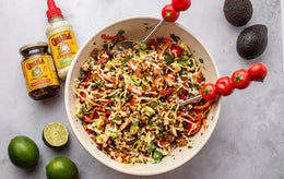 Salad in a bowl with ingredients including limes, avocados, and a jar of Cholula sauce on a gray surface.