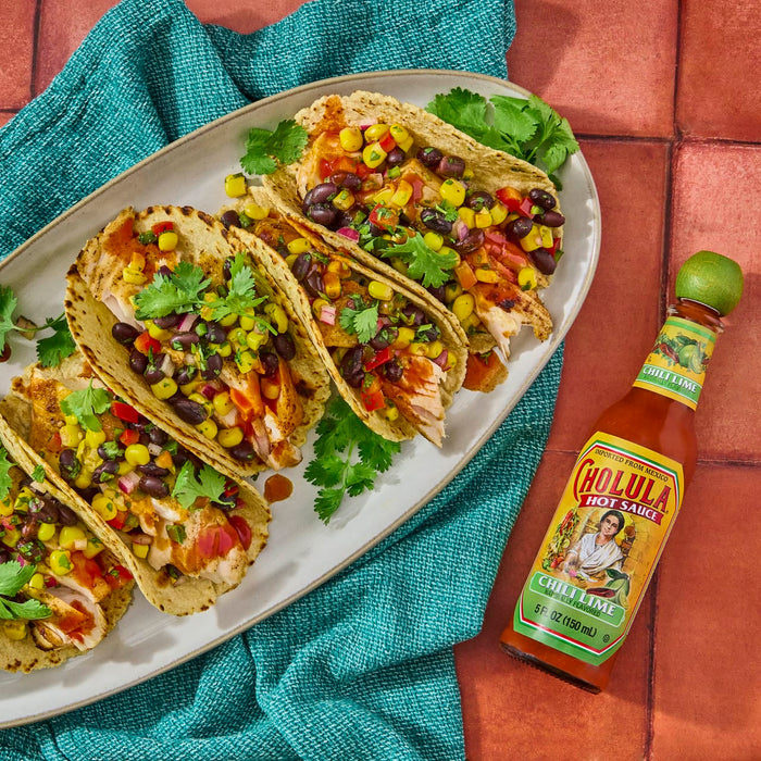 A plate of fish tacos with corn black beans and cilantro next to a bottle of Cholula Chili Lime hot sauce.