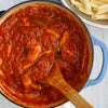 A pot of tomato sauce with chicken and peppers, a wooden spoon, and a strainer of cooked pasta in the background.
