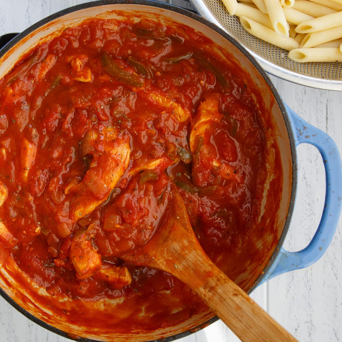 A pot of tomato sauce with chicken and peppers, a wooden spoon, and a strainer of cooked pasta in the background.