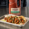 A gallon jug of Cattlemens BBQ Sauce Hawaiian Tangy Sweet sits next to a tray of three tacos.