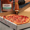 A hand in a black glove is squeezing Cattlemens BBQ sauce onto a Hawaiian pizza in a pizza box.