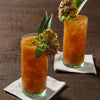 Two glasses of orange juice with pineapple and mint garnishes sit on white napkins on a wooden table.