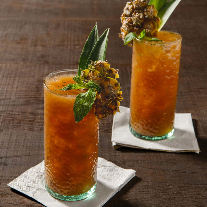 Two glasses of orange juice with pineapple and mint garnishes sit on white napkins on a wooden table.