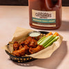 A bottle of Cattlemens BBQ Sauce Hawaiian Tangy Sweet with a basket of chicken wings, celery sticks, and carrots.