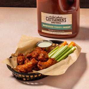 A bottle of Cattlemens BBQ Sauce Hawaiian Tangy Sweet with a basket of chicken wings, celery sticks, and carrots.