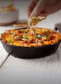 Hand dipping a chip into a skillet of nachos with cheeseburger dip and  green onions on a wooden surface.