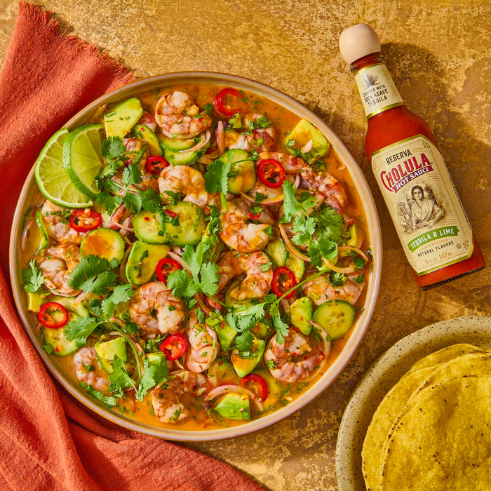 A bowl of shrimp ceviche with avocado, cucumber, red onion, cilantro, and red pepper with a bottle of Cholula Tequila &amp; Lime hot sauce and a plate of tortilla chips.