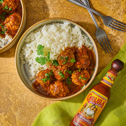 A bowl of white rice with meatballs in a spicy sauce and a bottle of Cholula Chipotle hot sauce.