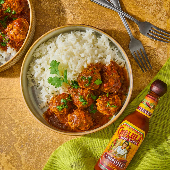 A bowl of white rice with meatballs in a spicy sauce and a bottle of Cholula Chipotle hot sauce.