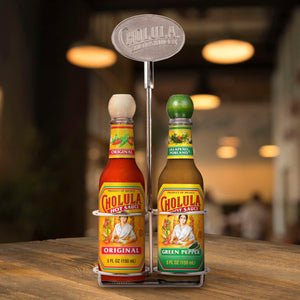 A metal tabletop caddy holds two five ounce bottles of Cholula Hot Sauce in Original and Green Pepper flavors against a blurred restaurant background.
