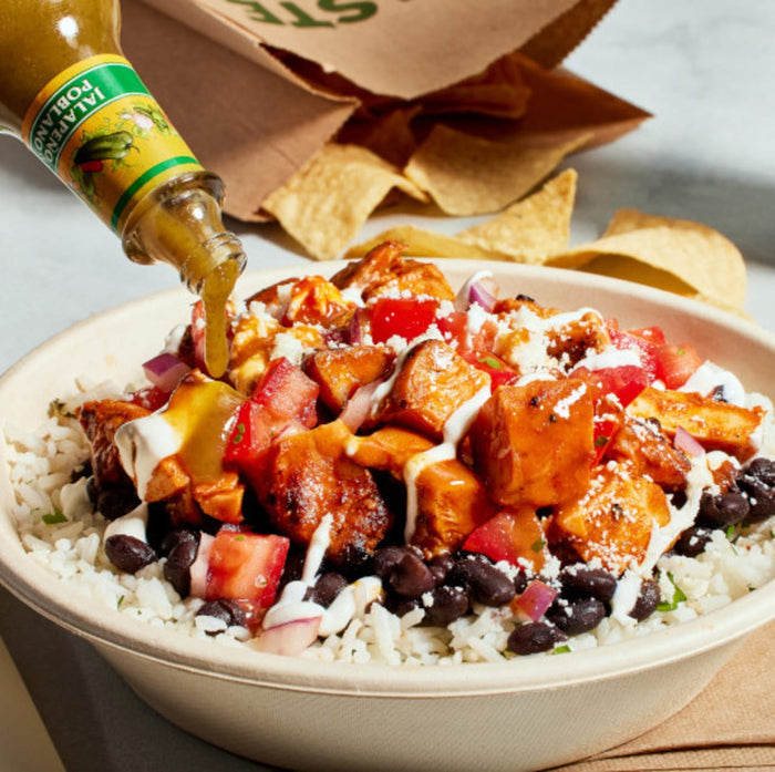 A bottle of Cholula Jalapeño Poblano Hot Sauce is being poured over a vibrant burrito bowl filled with white rice, black beans, grilled chicken, pico de gallo, and a drizzle of crema.