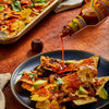 A bottle of Cholula Chipotle Hot Sauce is being poured over a plate of loaded nachos topped with melted cheese, beans, avocado, and cilantro.