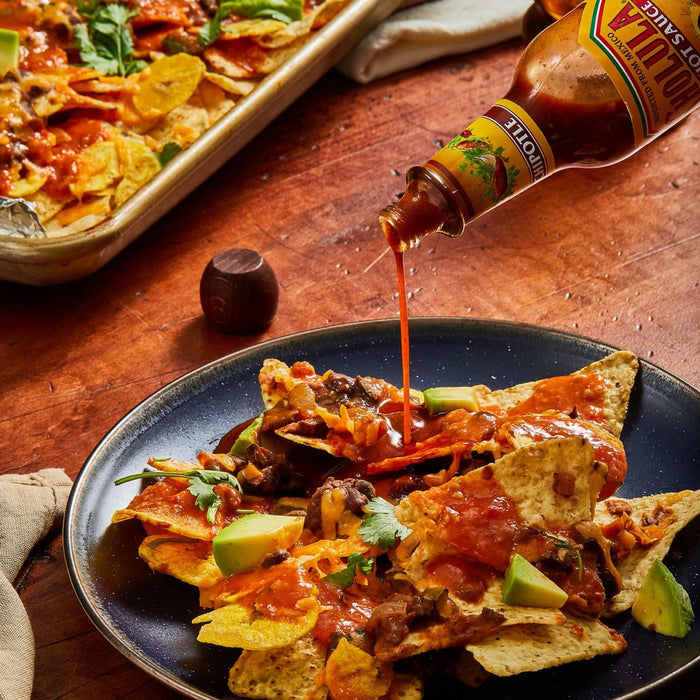 A bottle of Cholula Chipotle Hot Sauce is being poured over a plate of loaded nachos topped with melted cheese, beans, avocado, and cilantro.
