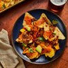 A blue plate holds a serving of loaded nachos topped with melted cheese, black beans, salsa, fresh avocado chunks, and cilantro sprigs next to a glass of beer.