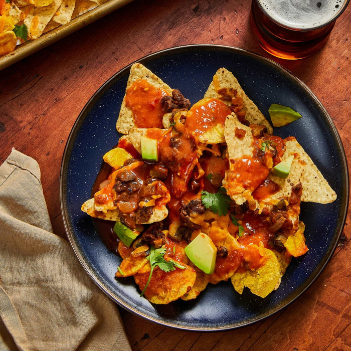 A blue plate holds a serving of loaded nachos topped with melted cheese, black beans, salsa, fresh avocado chunks, and cilantro sprigs next to a glass of beer.