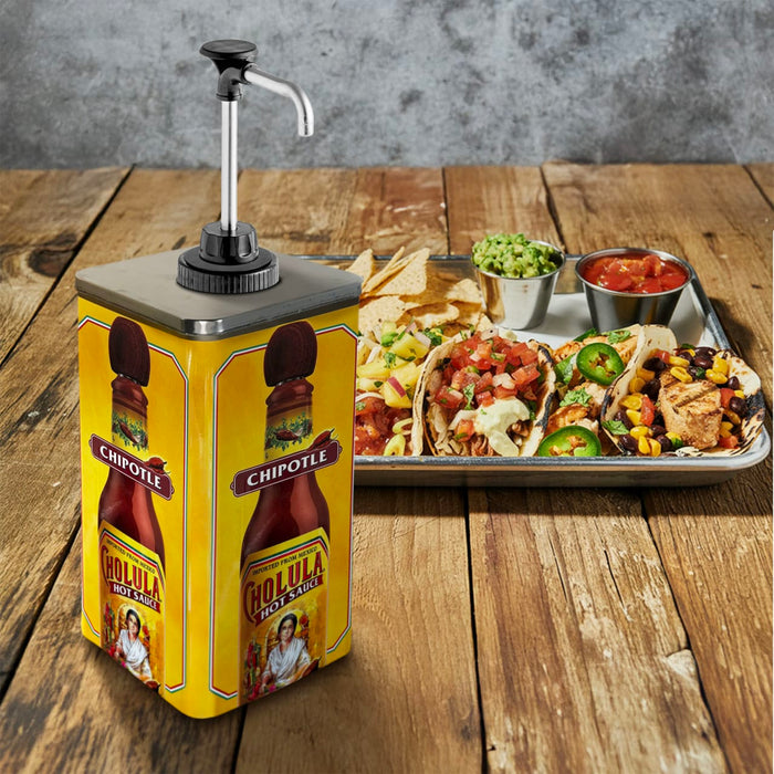 A large yellow dispenser of Cholula Chipotle Hot Sauce sits on a wooden table next to a tray of loaded tacos and chips with salsa and guacamole.