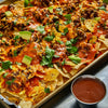 A sheet pan of loaded nachos topped with melted cheese, black beans, avocado chunks, and fresh cilantro is served alongside a small metal ramekin of red sauce.