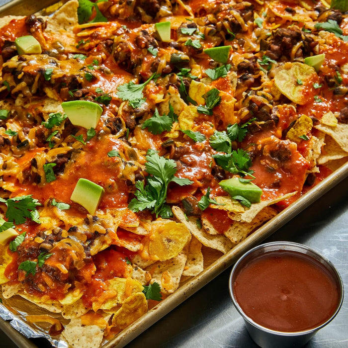 A sheet pan of loaded nachos topped with melted cheese, black beans, avocado chunks, and fresh cilantro is served alongside a small metal ramekin of red sauce.