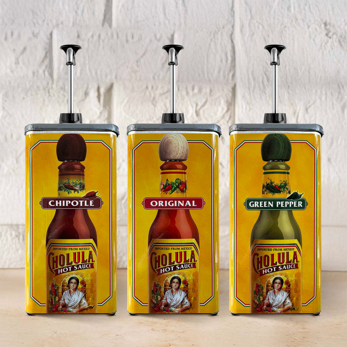 Three large yellow pump dispensers featuring the labels for Cholula Hot Sauce in Chipotle, Original, and Green Pepper varieties are lined up against a white brick wall.