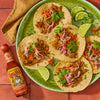 A plate of tacos with pulled pork, onions, cilantro, and lime wedges, with a bottle of Cholula Sweet Habanero hot sauce.