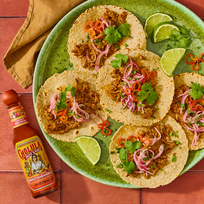 A plate of tacos with pulled pork, onions, cilantro, and lime wedges, with a bottle of Cholula Sweet Habanero hot sauce.