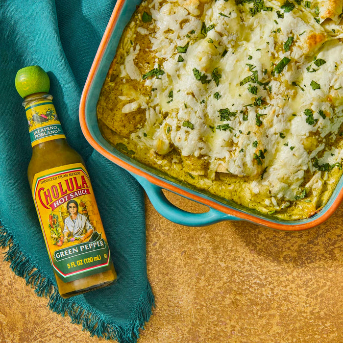 A bottle of Cholula Green Pepper hot sauce next to a dish of enchiladas.