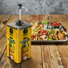 A large yellow dispenser of Cholula Green Pepper Hot Sauce sits on a rustic wooden table next to a tray of loaded tacos and chips.