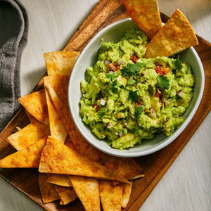 A bowl of chunky guacamole garnished with fresh cilantro and diced tomatoes sits on a wooden tray alongside a pile of seasoned tortilla chips.
