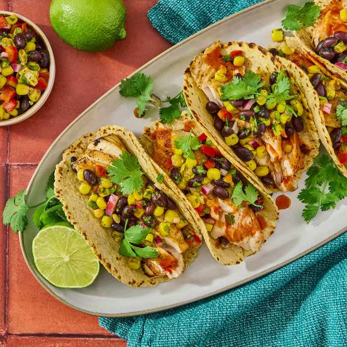 A platter of grilled chicken tacos topped with a vibrant corn and black bean salsa and fresh cilantro sprigs.